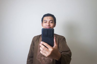 Cheerful young Asian Muslim man using a mobile phone and looking away isolated over white background clipart