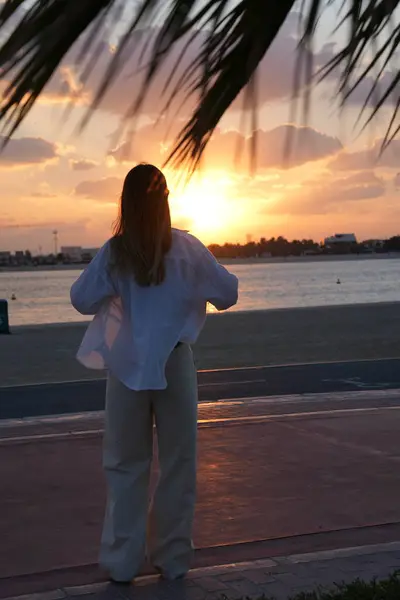 Gün batımını izleyen, tropik bir sahilde palmiye yapraklarının altında duran bir kadının silueti. Huzurlu bir yalnızlık, tropikal doğa ve tefekkür anı. Romantik, seyahat ve doğa temalı yayınlar için harika.
