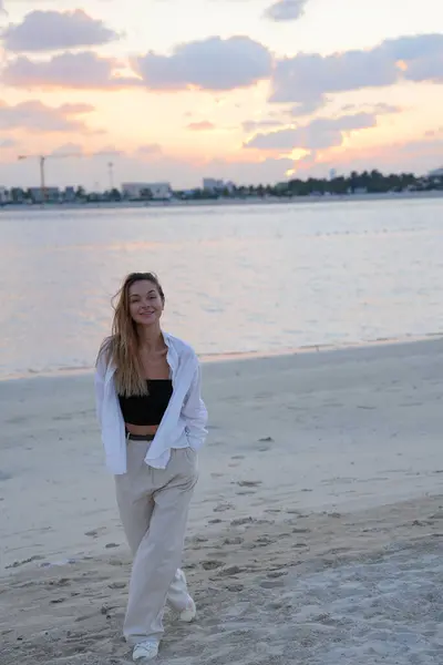 Kumlu bir sahilde hafif günlük giysiler içinde yürüyen neşeli genç bir kadın. Yaşam tarzı blogları, yaz reklamları ve seyahat promosyonları için uygun.