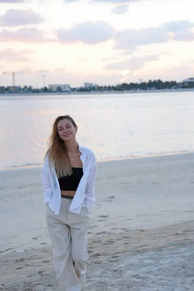 Beyaz pantolonlu ve gömlekli genç bir kadın gün batımında denizin kenarında dikiliyor. Barış, farkındalık, moda ya da yaşam tarzı kampanyaları için idealdir..