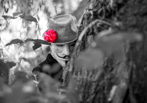 Close-up portrait of a masked man in a hat and a rose on his head against the background of a summer forest and a tree. Black and white photo