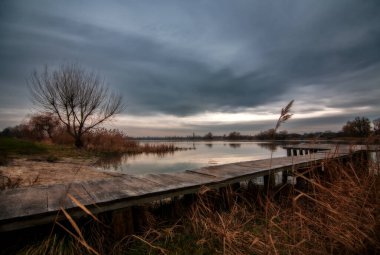 Akşam gökyüzüne karşı kış manzarası, nehrin yanında yapayalnız bir ağaç, kara bulutlar ve kuru otlardan oluşan bir ada. Gökyüzü farklı renklerde ve su yaprakları olmayan bir ağacı yansıtıyor. Önplanda uzun bir köprü.