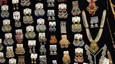 Renkli etnik küpe ve kolyelerin detaylı görünümü siyah bir arkaplanda düzenlenmiştir. Çeşitli stil ve tasarımlar arasında inci süslemeler ve karmaşık metal işçiliği yer almaktadır..