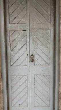 Açık gri boyalı, köşegen destekleyici, çivili inşaat ve merkezden sarkan paslı kilitlerin yer aldığı eski bir çift kapılı yakın plan..