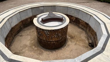 Eski bir açık hava çeşmesinin detaylı yüksek açılı çekimi. İçinde gri aksanlı mermer duvarlar, kahverengi metal bir süs ve yapıyı çevreleyen taş bir zemin var..