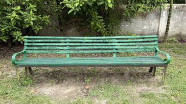Yeşillik bir park bankının manzarası ahşap çıta ve metal çerçeve ile yemyeşil ağaç yaprakları altında, bir bahçe arka planında yıpranmış beton bir duvar karşı.