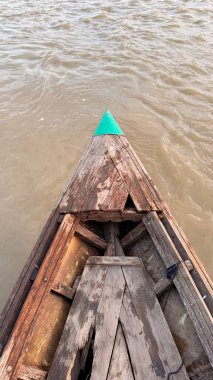 Göz hizasında çekimde, çamurlu bir nehri kesen eski, ahşap bir teknenin karmaşık dokuları, belirgin yeşil üçgen yayı ve parlak bir kontrastı gösteriyor..