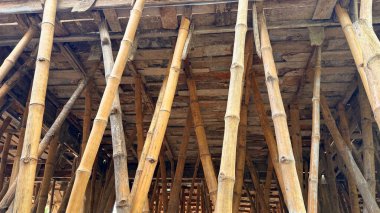 Bu düşük açılı çekim, açık kahverengi bambu direklerden oluşan çetrefilli ağı yakalar ve yukarıdaki aşınmış ahşap iskeleti destekleyen yapısal rollerini gösterir. Görüntü ham madde ve yapıyı gösteriyor.