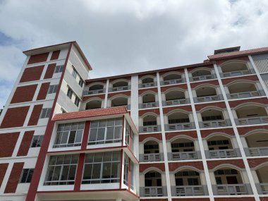 Aşağıdaki manzara, bulutlu ve bulutlu bir gökyüzünün arka planına vurgulanmış, beyaz aksanlı kırmızı tuğlalı bir cepheye sahip çok katlı bir binayı gösteriyor..