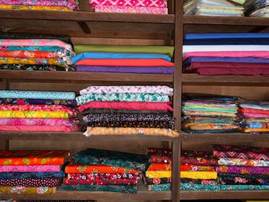 Ahşap raflar düzgün desenli ve sağlam tekstil sergiliyor, düzenli bir şekilde istiflenmiş, çeşitli dizayn ve renklerde katlanmış kumaşların çeşitli bir koleksiyonunu sergiliyor, bir dizi tekstil seçeneği sunuyor..