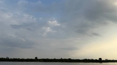 Sakin bir nehir, gün batımında, ağaçlarla benekli yeşil bir nehir kıyısında, bulutlu bir gökyüzünü yansıtır. Huzurlu manzara rahatlatıcı bir manzara yaratır..