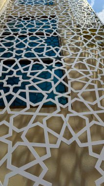 Beyaz geometrik kafes desenleri, pencereleri düşük açıdan gören bir binanın ön cephesini süslüyor. Kamp çalışması binanın yüzeyinde karmaşık gölgeler yaratıyor ve açıklıklardan mavi cam parçaları geçiyor. Yüksek çözünürlüklü fotoğraf.