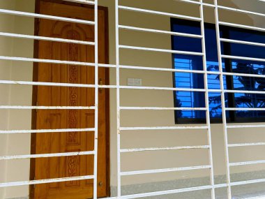 Yüksek çözünürlüklü bir fotoğraf, dış tarafta süslü ahşap bir kapı ve bir evin renkli camlı penceresini koruyan rustik beyaz bir metal parmaklık gösteriyor..