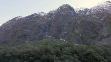 Yeni Zelanda 'daki Milford Sound' a giderken birçok şelalesi olan bir yamaç. Tepelerin tepelerinde kar var ve bir sürü yeşil yaşam da.