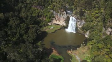 Auckland, Yeni Zelanda 'daki Hunua Falls adlı bir şelalenin insansız hava aracı görüntüsü. Şelale ormanla çevrilidir. Atış yukarıdan yapılıyor ve dönüyor.