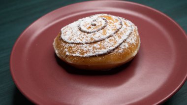 Üzerinde pudra şekeri ve yuvarlak çikolata sosu olan taze bir çörek, kırmızı tabakta servis edilir..