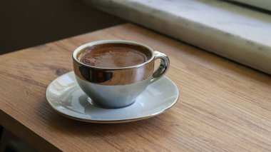 Köpüklü kahveyle dolu parlak gri bir kahve fincanı, arka plandaki cam pencere duvarına bağlı ahşap bir tahta, güpegündüz çekilmiş..