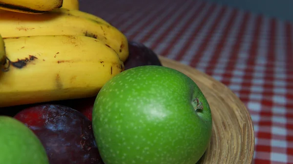 a close-up of a fruit plate on a wooden plate consisting of a green apple, a banana and a large plum