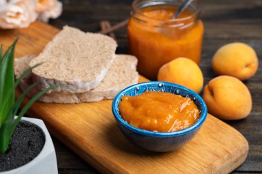 Mavi bir seramik kasede kayısı reçeli, ahşap zemin üzerinde ekmek ve taze kayısılar.