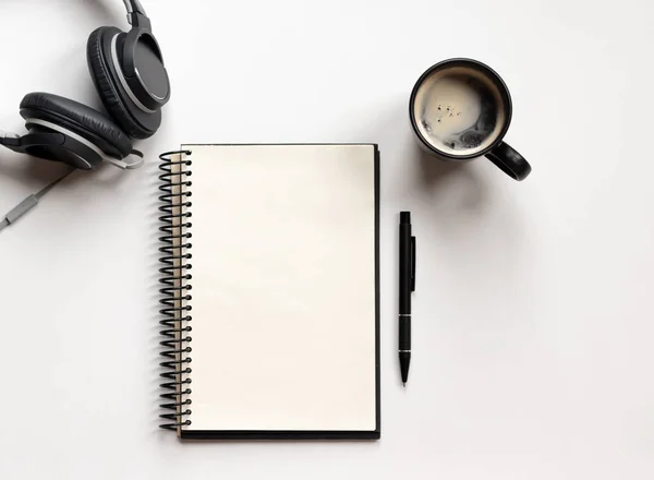 Modern white office desk table with notebook, pen, headphone and coffee cup. Blank notebook page for text entry in the middle. Top view, flat lay.