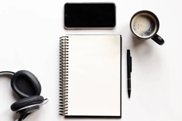 Modern white office desk table with notebook, pen, smartphone, headphone and coffee cup. Blank notebook page for text entry in the middle. Top view, flat lay.