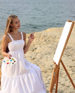 Yaratıcı kadın sanatçı, doğada yeni bir fikir tablosu üzerinde çalışıyor. O, boya, kalem, palet ve bir fırçayla resim çiziyor. O, denizin yanında oturuyor. Gülümsüyor.. 