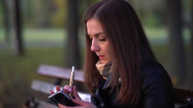 Genç bir kadının dışarıda elektronik sigara içerken 4K pozu. Kadın elektronik sigara buharı soluyor ve sonbahar parkında telefonuna mesaj yazıyor. Teknoloji kullanılıyor