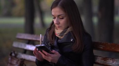 Genç bir kadının dışarıda elektronik sigara içerken 4K pozu. Kadın elektronik sigara buharı soluyor ve sonbahar parkında telefonuna mesaj yazıyor. Teknoloji kullanılıyor