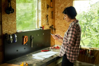 Female business owner using digital tablet in workshop, industrial woman engineer working with blueprints and tablet in the workshop.