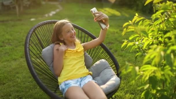 幸せな子供の女の子は公園の屋外で携帯電話で自撮り 子供はスマートフォンを使用して自宅の庭 日光で写真を撮る — ストック動画