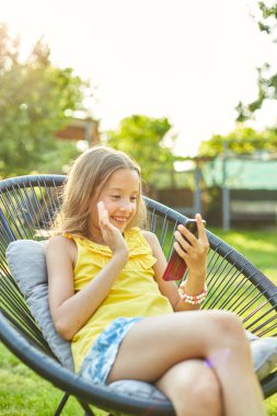 Happy kid girl holding smartphone having video call with friend distantly, online talk in the park outdoor, child using smartphone at home garden, backyard, sunlight, communication, technology concept