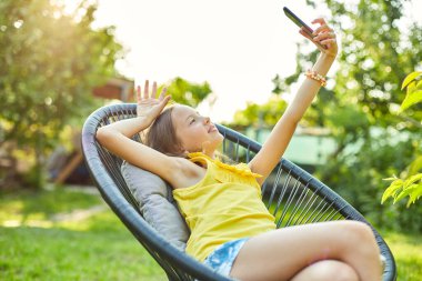 Mutlu çocuk parkta cep telefonuyla selfie çekiyor, akıllı telefon kullanarak ev bahçesinde, arka bahçede, gün ışığında fotoğraf çekiyor.