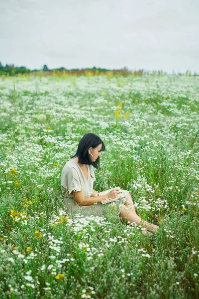 Female woman with pen writing or painting, handwriting on notebook on flower blooming meadow, in countryside outdoor, in the field on summer day in meadow, feel the nature, relax, alone travel.