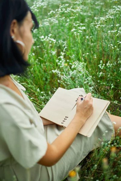 Female woman with pen writing or painting, handwriting on notebook on flower blooming meadow, in countryside outdoor, in the field on summer day in meadow, feel the nature, relax, alone travel.