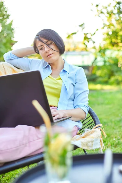 Freelance woman works on laptop at home in the backyard, Concept of remote work from comfortable outdoor workspace, checking social media, shopping online, ordering delivery in the garden.