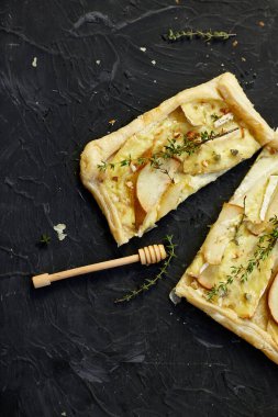 Pie baked in puff pastry with gorgonzola cheese and pears on black table, cutting pizza with pear, thyme, copy space, top view.