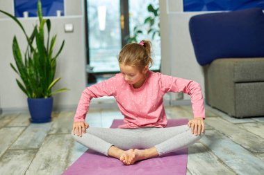 Küçük kız esneme egzersizleri yapıyor, evde fitness minderinde yoga yapıyor spor yapıyor, sağlıklı bir yaşam tarzı sergiliyor. Çocuklar ev karantinası yapıyor
