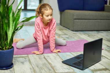 Küçük kız dizüstü bilgisayarda video izliyor ve evde esneme hareketleri yapıyor. Kişisel antrenörle uzak çocuk eğitimi, online eğitim konsepti, spor yapmak, sağlıklı yaşam tarzı.