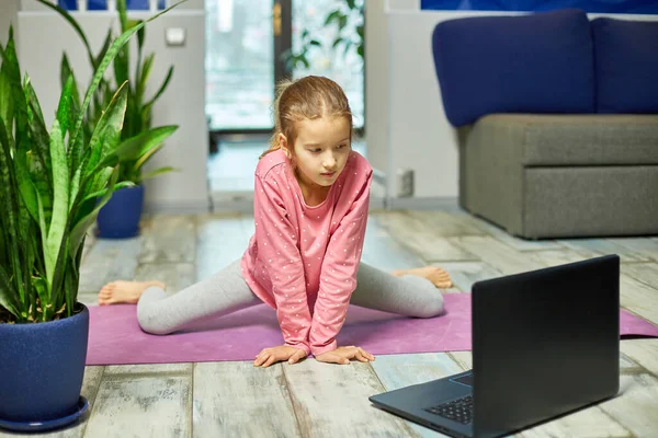 Little girl watching online video on laptop and doing stretching, fitness exercises at home. Child distant training with personal trainer, online education concept, Playing sports, Healthy lifestyle.