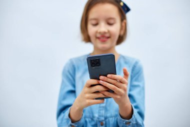 Smiling little girl in casual denim dress hold smartphone, using mobile cell phone internet applications, typing message isolated on white background, studio. Child and modern tech usage concept