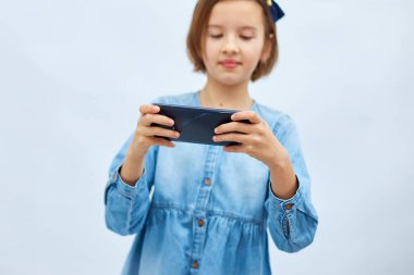 Sad kid girl in casual denim dress use with smartphone, enjoys mobile app, internet addiction, play app on mobile cell phone hold gadget isolated on white background, studio