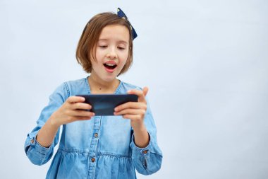 Fun excited kid girl in casual denim dress use with smartphone, enjoys mobile app, internet addiction, play app on mobile cell phone hold gadget isolated on white background, studio