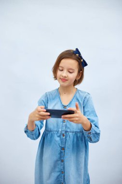 Fun excited kid girl in casual denim dress use with smartphone, enjoys mobile app, internet addiction, play app on mobile cell phone hold gadget isolated on white background, studio