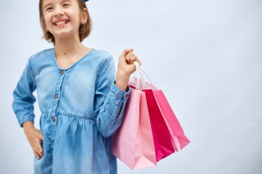 Pretty child girl in casual denim dress hold in hand pink shopping bags on white background, studio, Shopaholic shopping and fashion, discount sale concept, copy space, banner. Black friday