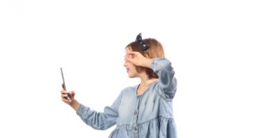 Smiling little girl in casual denim dress hold smartphone do selfie shot on mobile cell phone isolated on white background, studio. lifestyle concept.