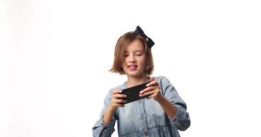 Fun excited kid girl in casual denim dress use with smartphone, enjoys mobile app, internet addiction, play app on mobile cell phone hold gadget isolated on white background, studio