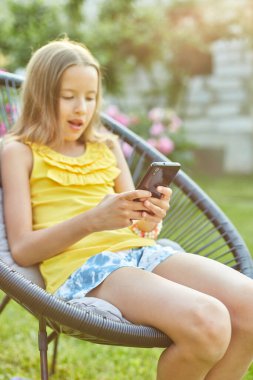 Parkta cep telefonuyla oyun oynayan mutlu bir kız çocuğu ev bahçesinde, arka bahçede akıllı telefon kullanan bir çocuk, güneş ışığı, akıllı telefon bağımlılığı.