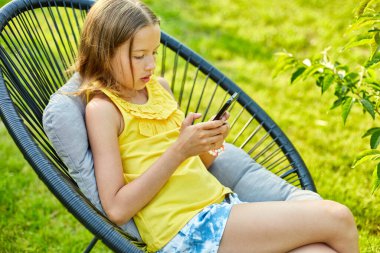 Parkta cep telefonuyla oyun oynayan mutlu bir kız çocuğu ev bahçesinde, arka bahçede akıllı telefon kullanan bir çocuk, güneş ışığı, akıllı telefon bağımlılığı.