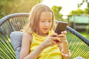 Parkta cep telefonuyla oyun oynayan mutlu bir kız çocuğu ev bahçesinde, arka bahçede akıllı telefon kullanan bir çocuk, güneş ışığı, akıllı telefon bağımlılığı.