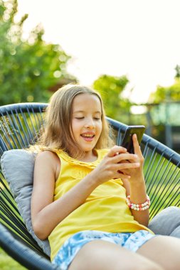 Parkta cep telefonuyla oyun oynayan mutlu bir kız çocuğu ev bahçesinde, arka bahçede akıllı telefon kullanan bir çocuk, güneş ışığı, akıllı telefon bağımlılığı.